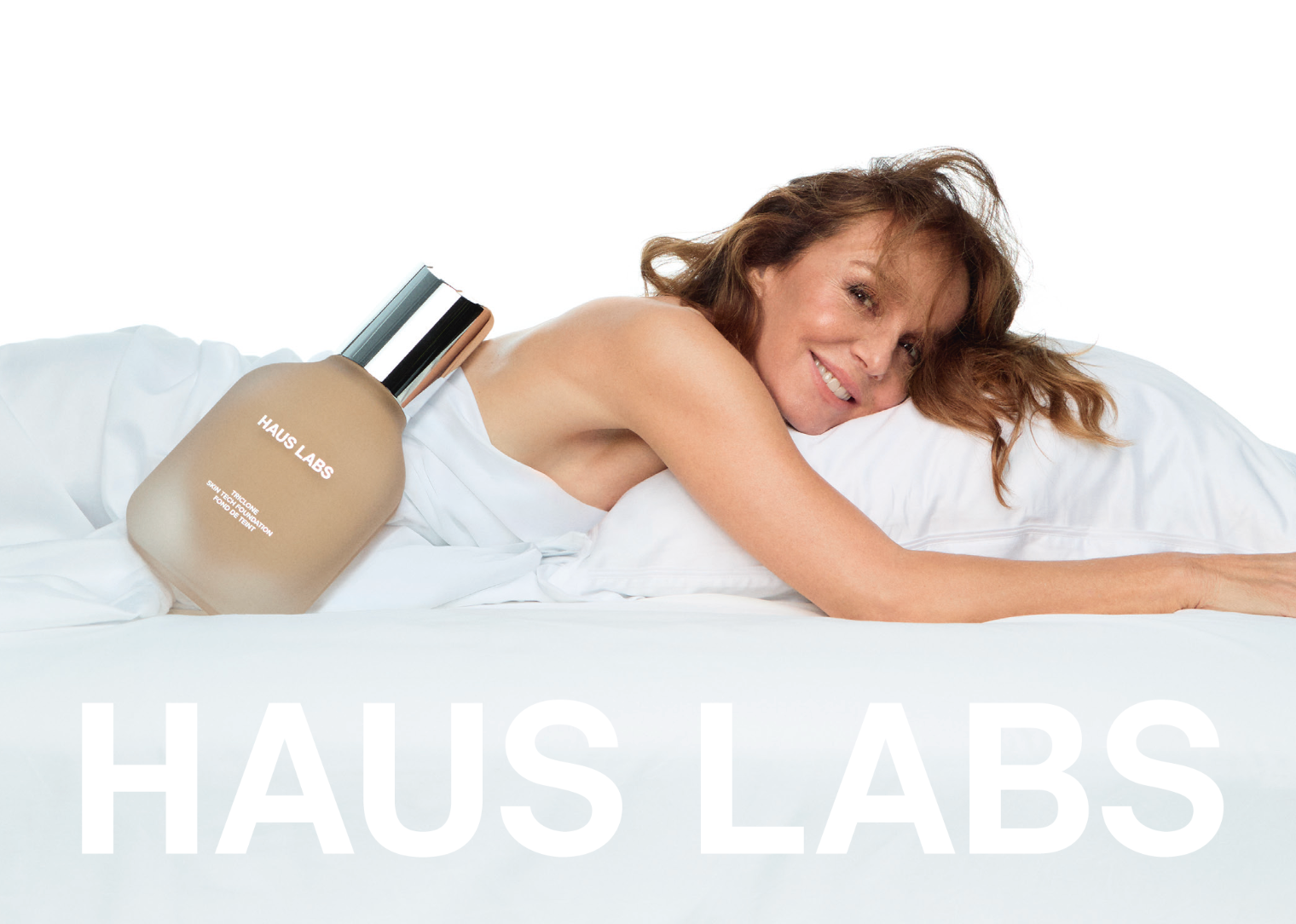 Woman with brown wavy hair lying on white bedding, smiling at camera while holding beige liquid foundation bottle with silver cap labeled HAUS LABS