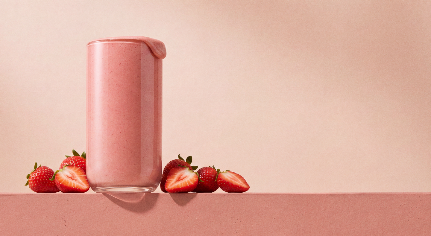 Glass of pink strawberry shake with fresh strawberries arranged around it on pink surface