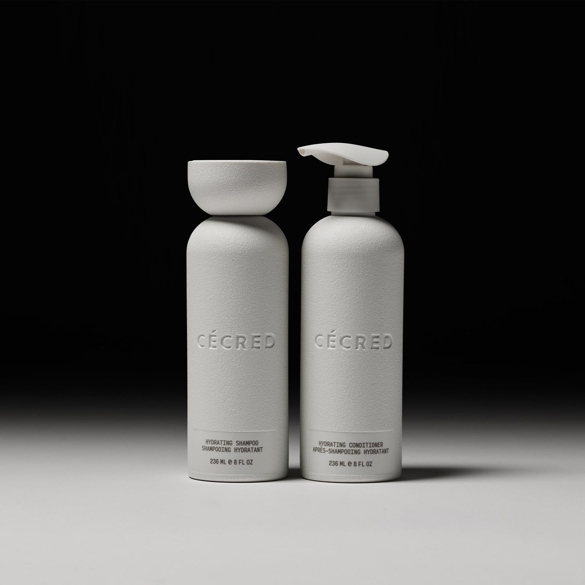 Two white cylindrical bottles labeled with product names and descriptions, one with a pump dispenser, on a gray surface