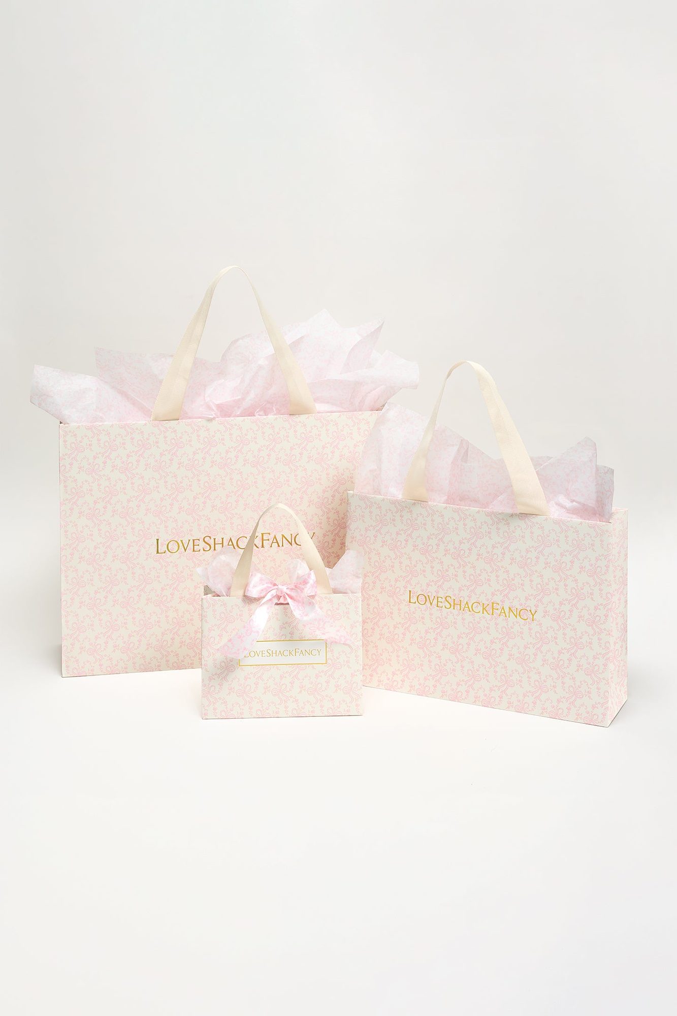 Three sizes of cream-colored shopping bags with tissue paper and ribbon bows