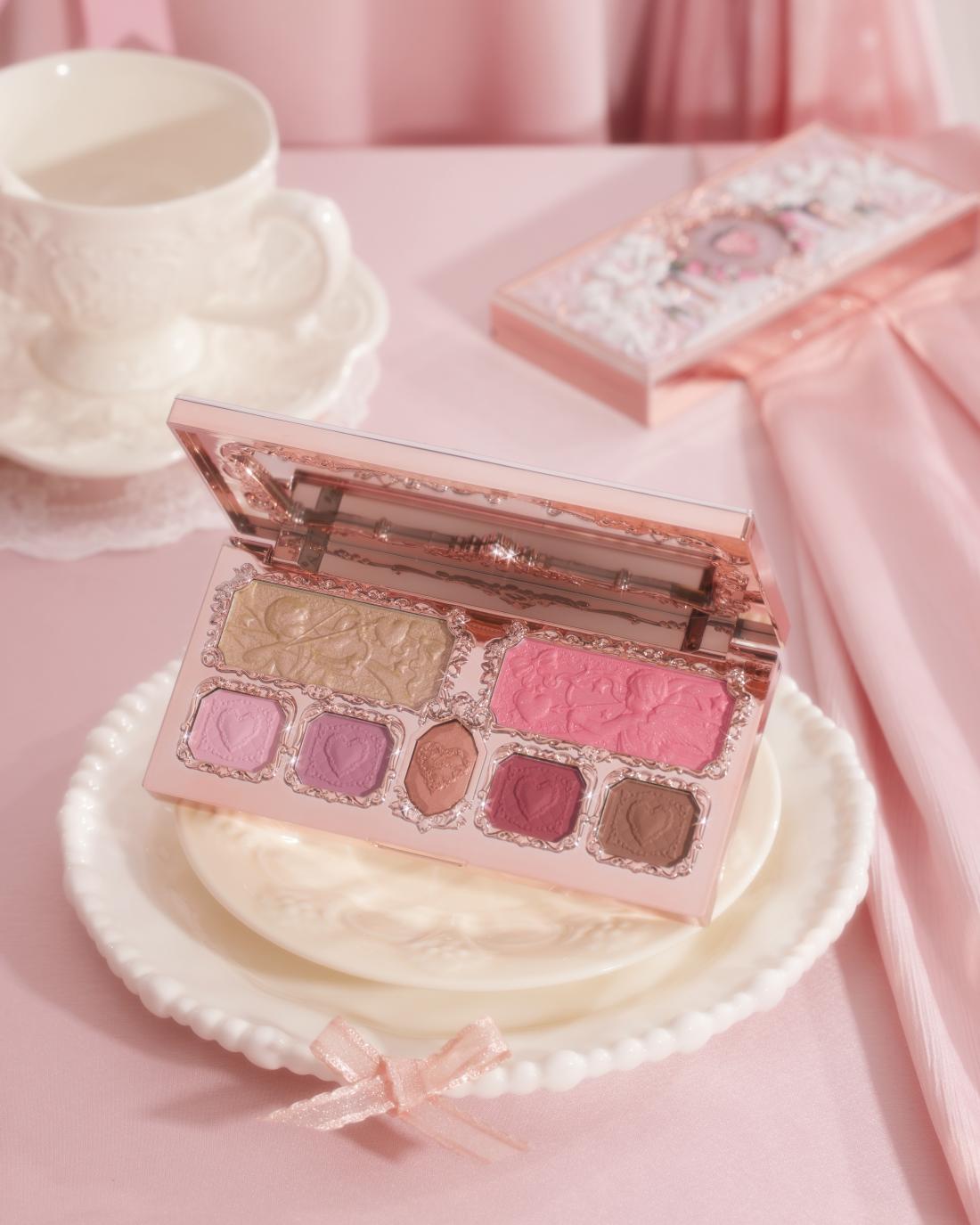 Pink makeup palette with multiple eyeshadow and blush shades displayed on white plate with coffee cup