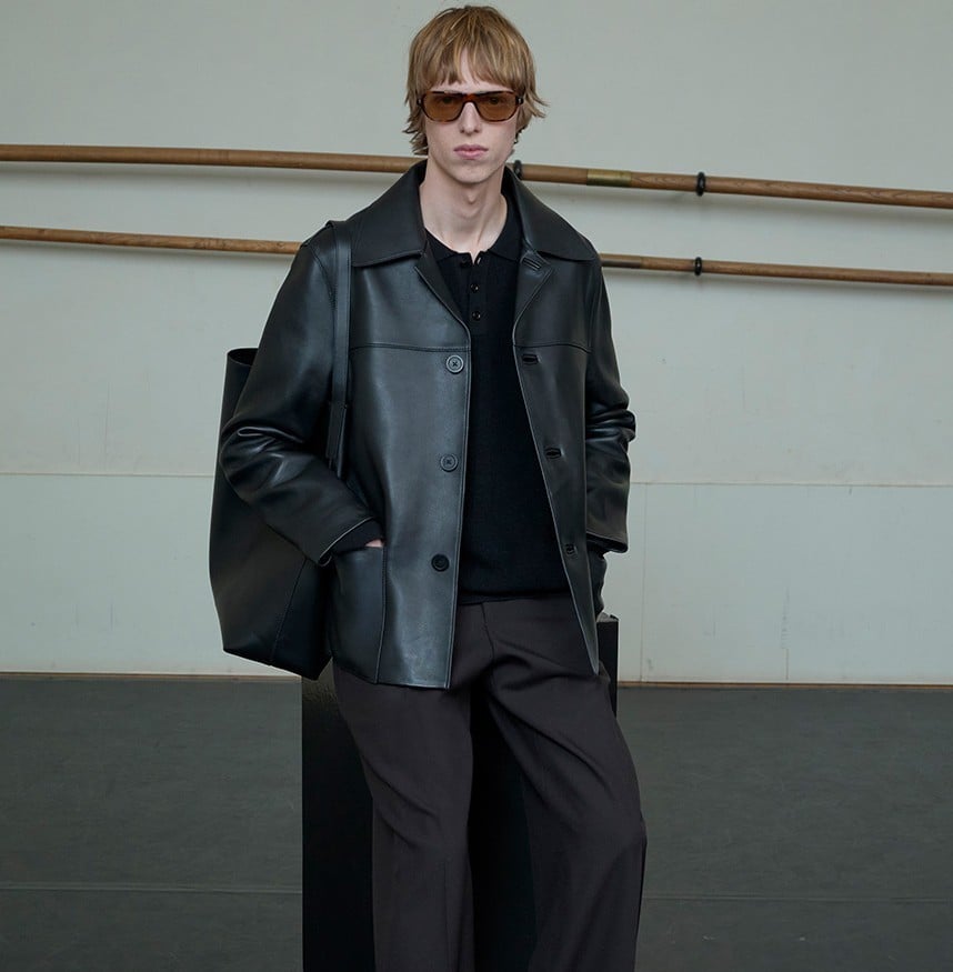 Person in black leather jacket and dark pants wearing brown-tinted sunglasses against white wall