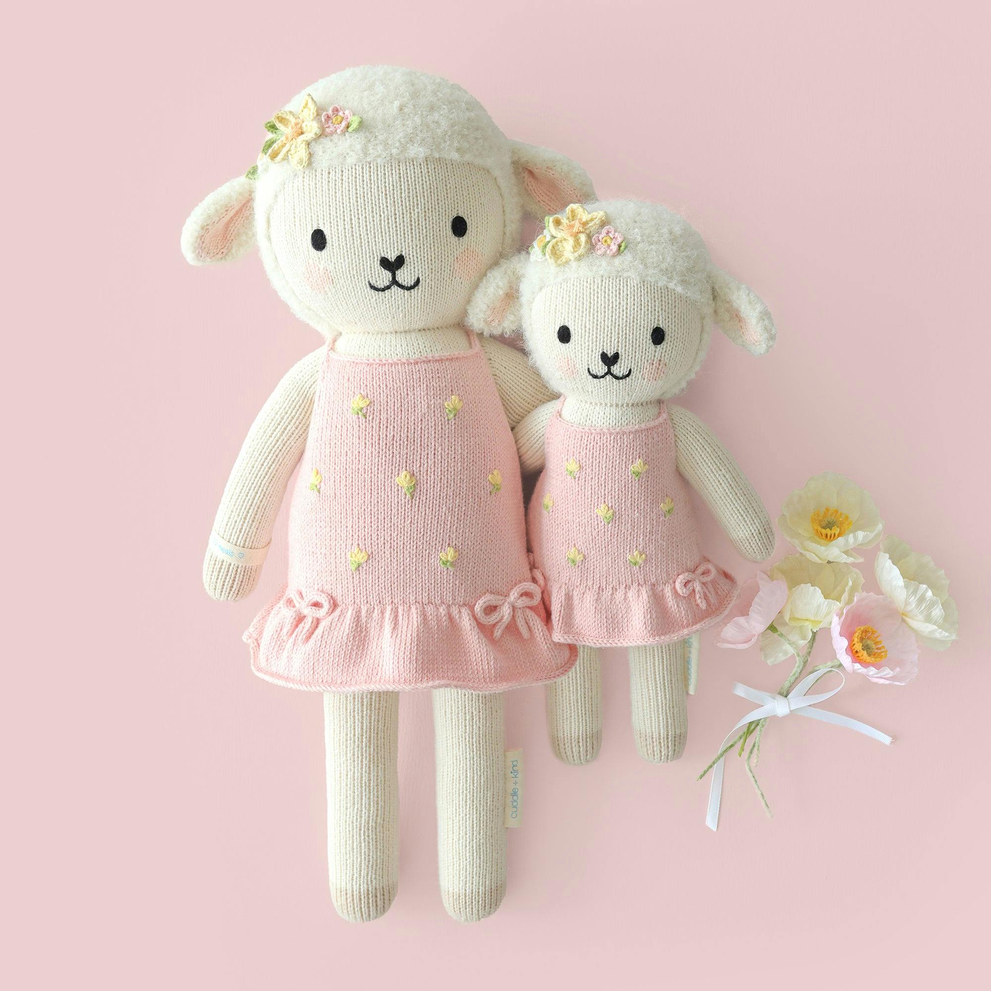 Two cream knit lamb dolls wearing pink dresses with yellow flower hair clips and holding white flowers