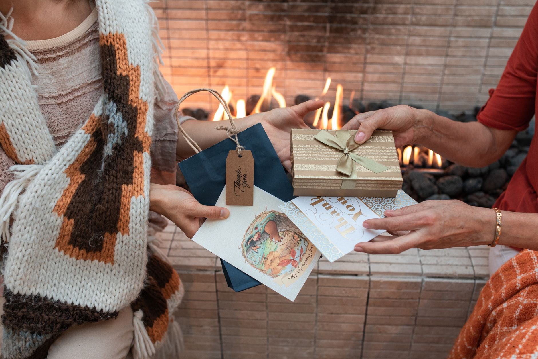 Two people in cozy sweaters exchanging wrapped gifts and greeting cards by a fireplace outdoors