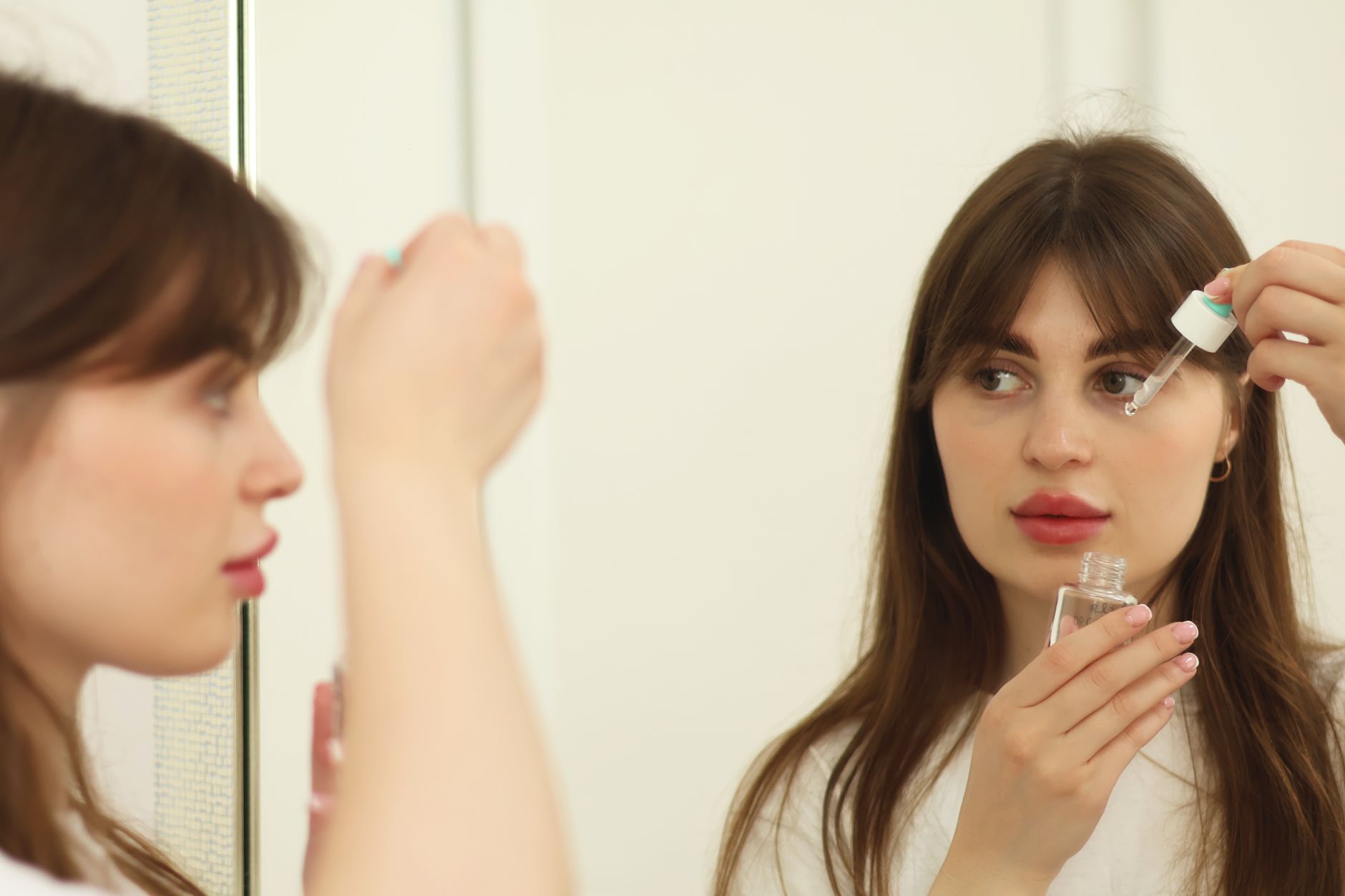 Woman applying facial serum drops to under-eye area while looking in mirror