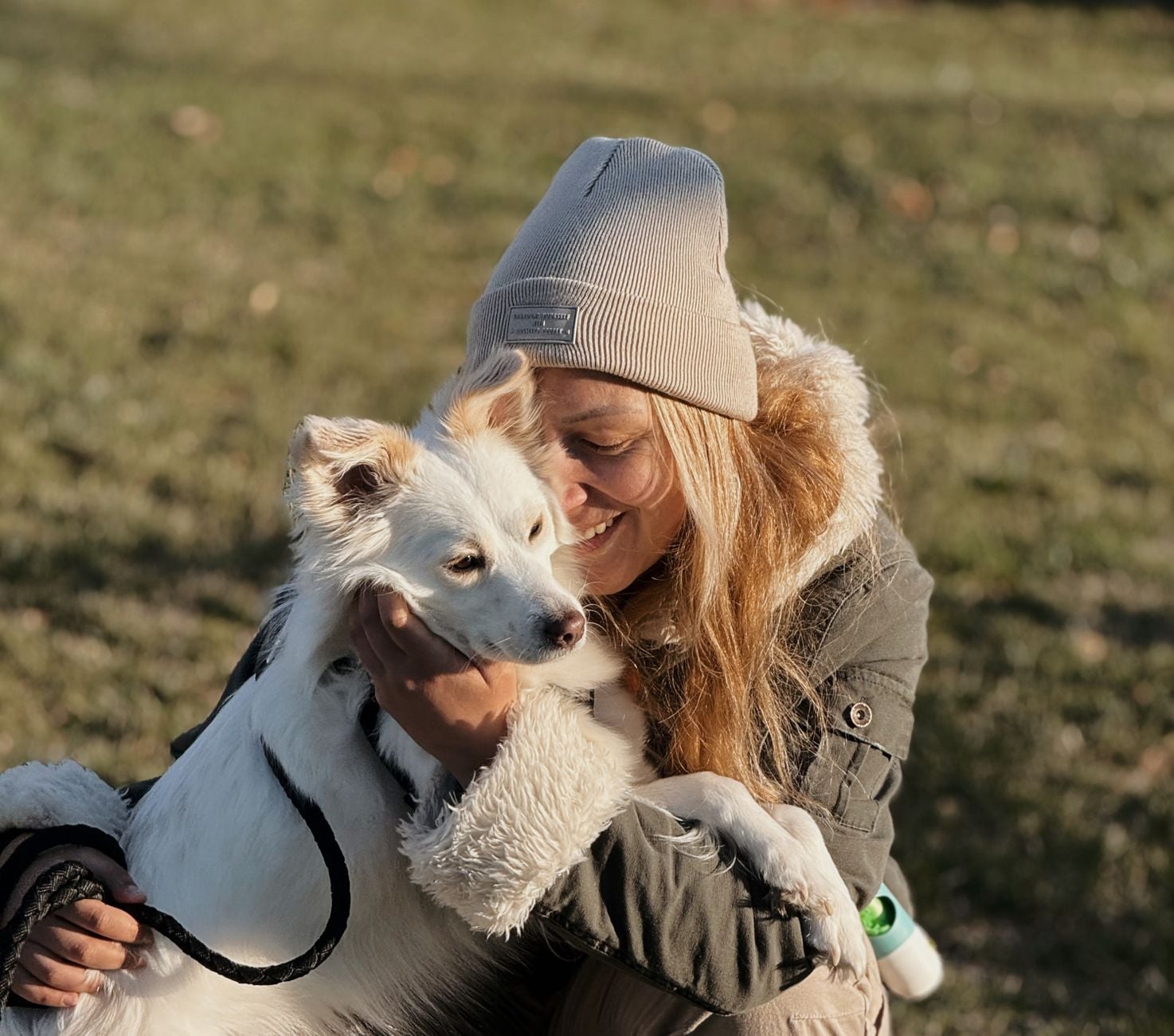 Woman in light blue beanie holding and smiling at a white dog outdoors on a grassy field