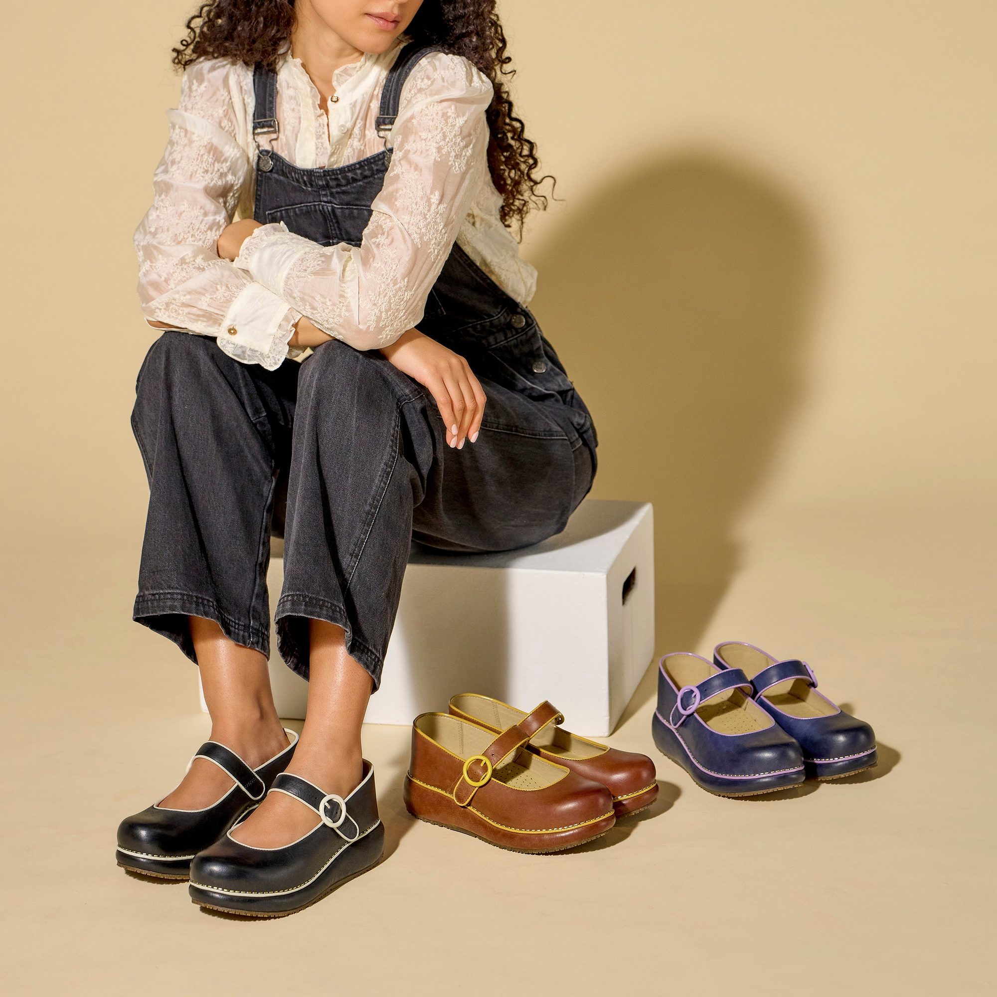 Person wearing black overalls with white lace shirt, displaying three different colored platform clogs in brown, tan, and navy