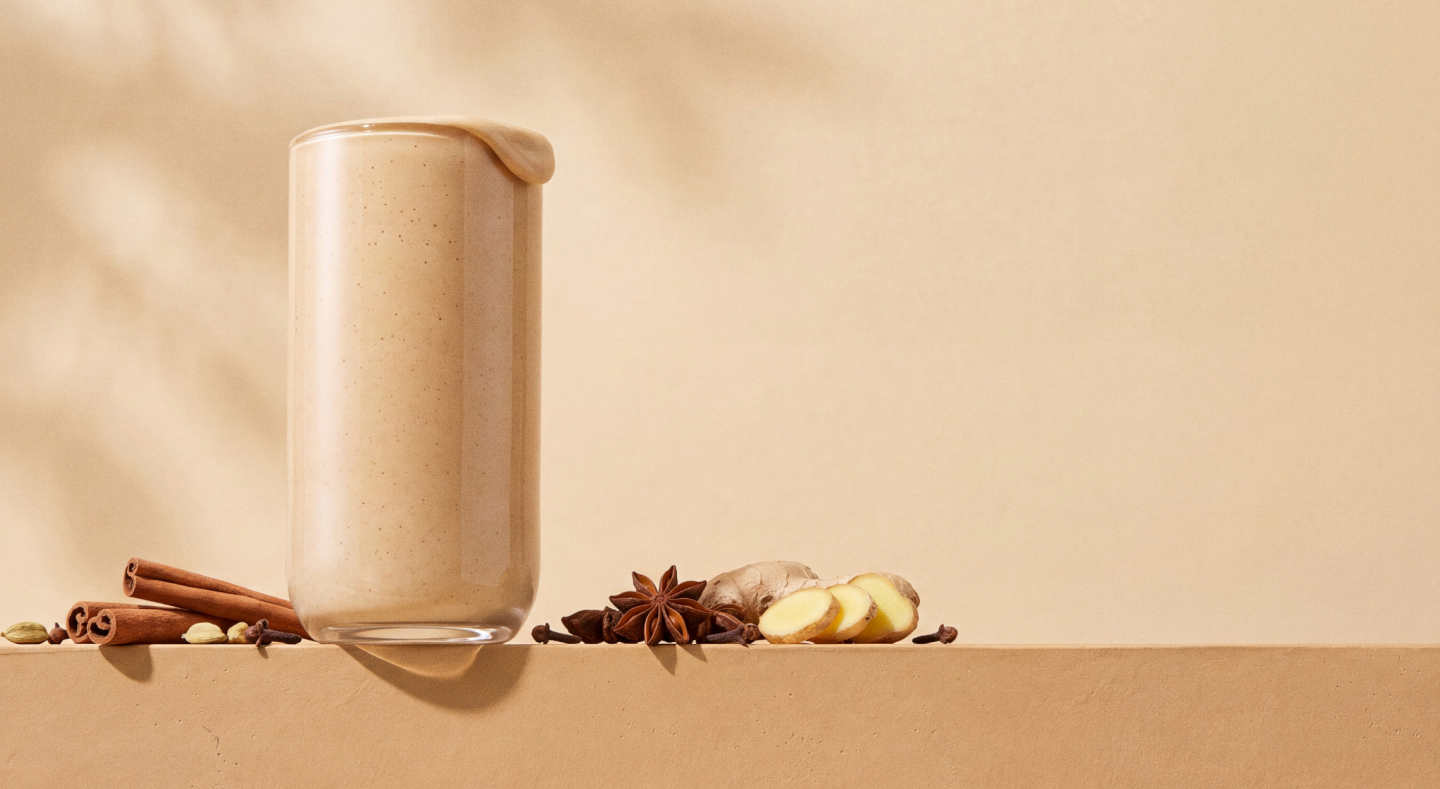 Glass of beige spiced shake surrounded by cinnamon sticks, star anise, and ginger slices