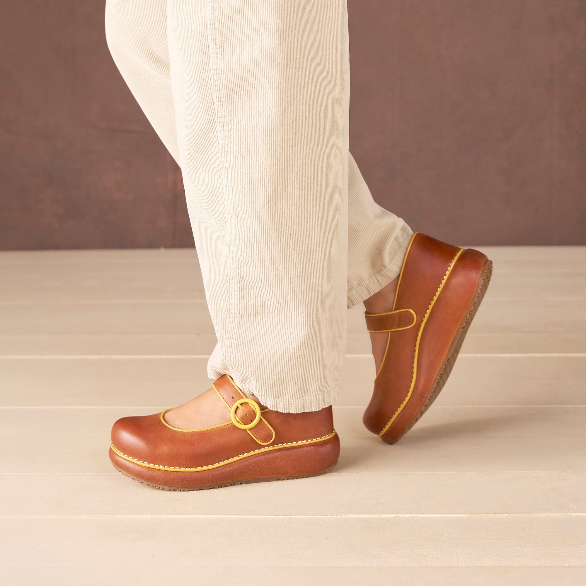 Brown leather platform Mary Jane clog with yellow stitching and buckle strap, worn with cream ribbed pants