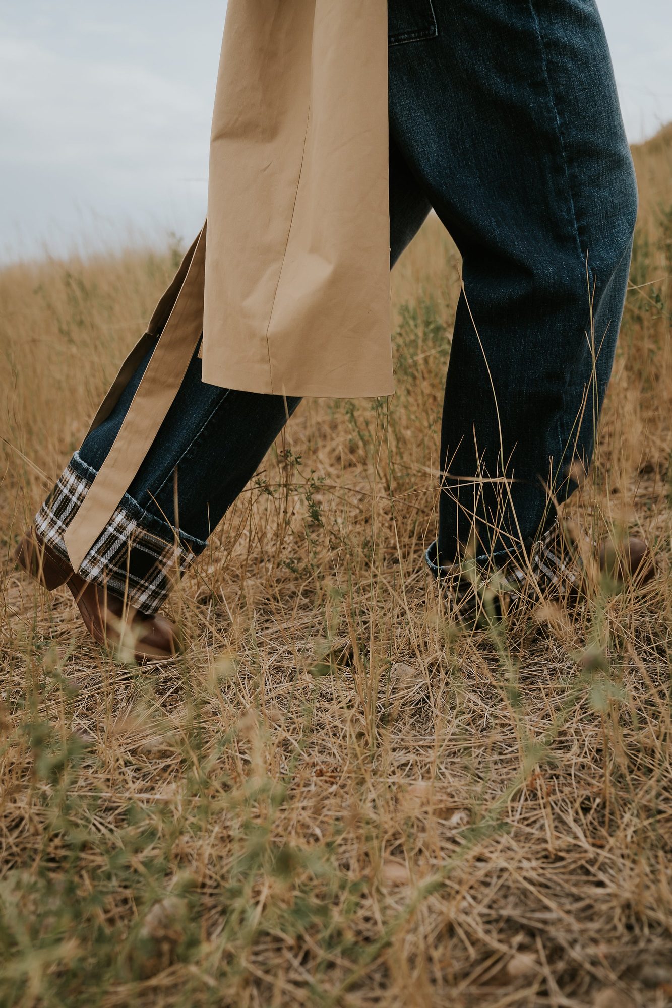 Close-up of woman's lower half showing beige coat, dark jeans, and plaid cuff details standing in field
