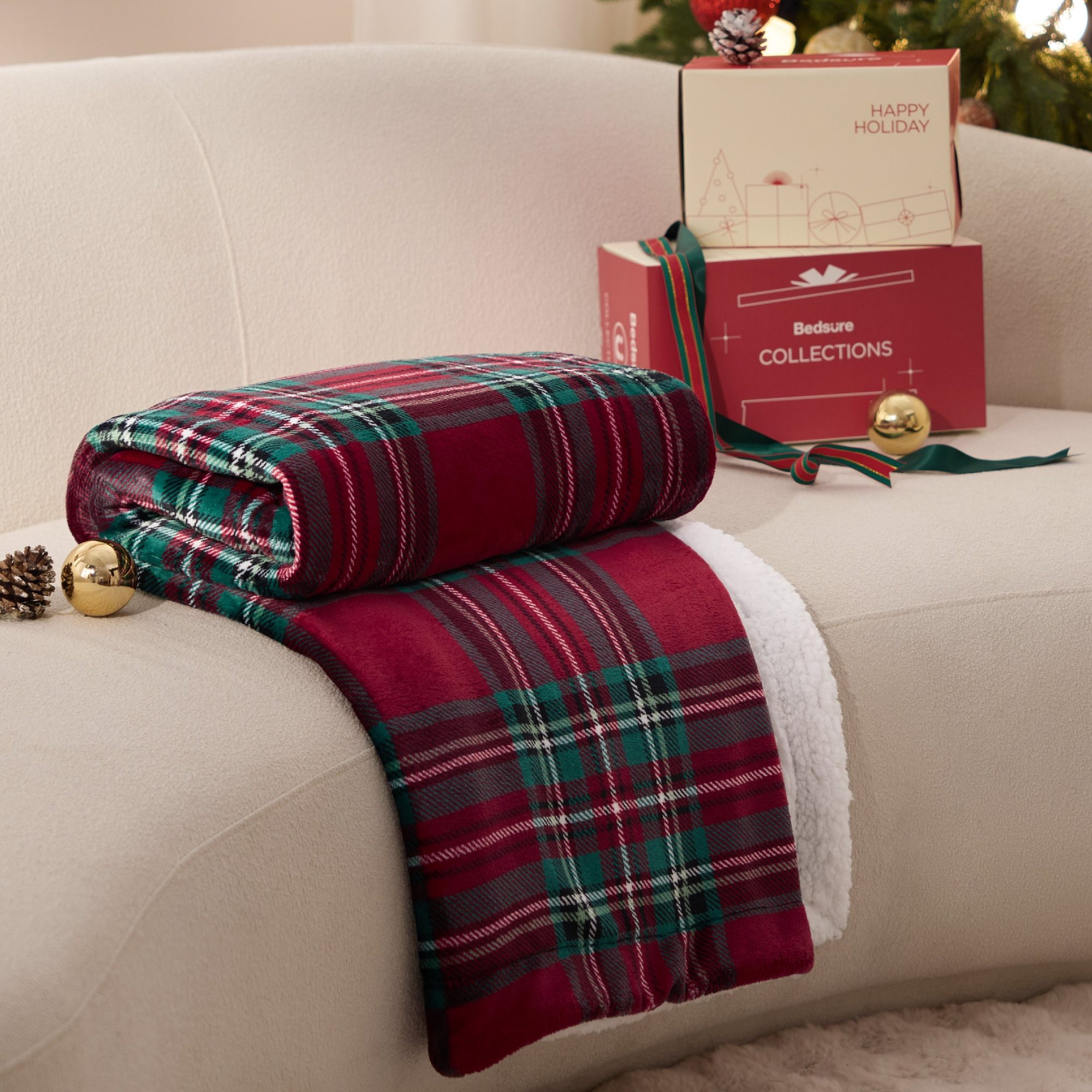 Red and green plaid blanket rolled up next to gift boxes on cream couch with holiday decorations
