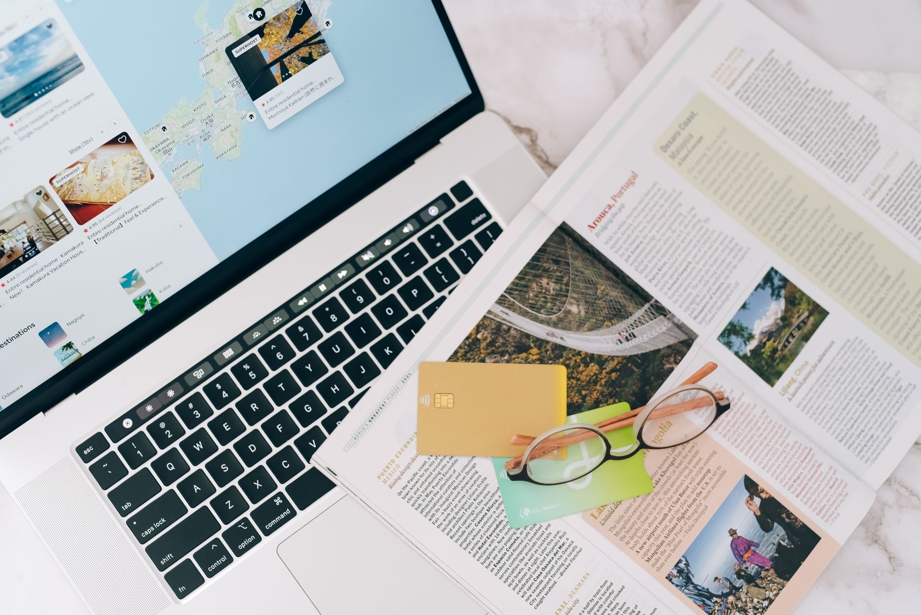 Laptop with travel website displayed, surrounded by colorful credit cards, eyeglasses, and travel magazine pages with destination photos