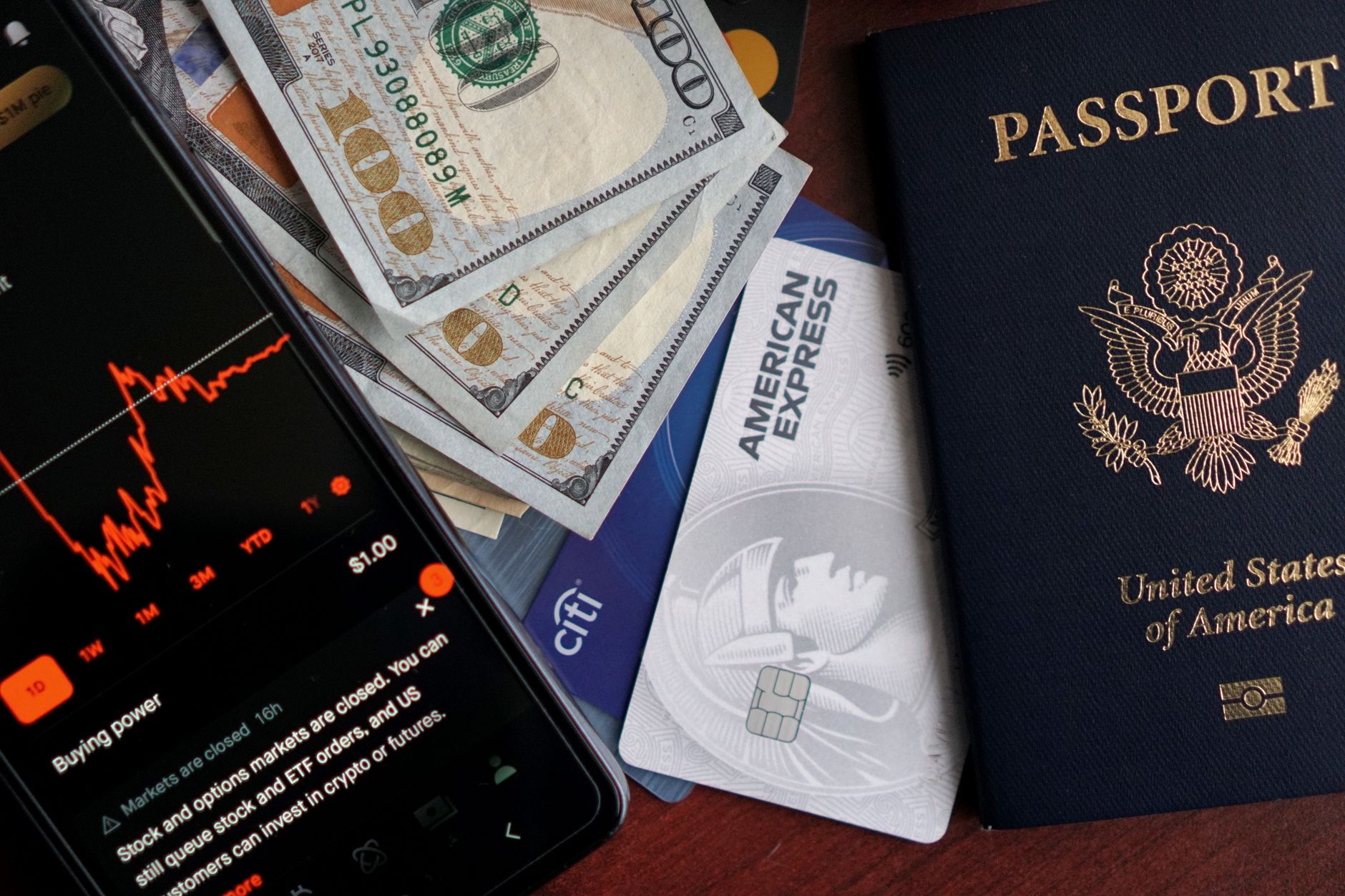 US passport and American Express traveler's checks next to US dollar bills, with a smartphone displaying a stock trading app showing a red downward chart