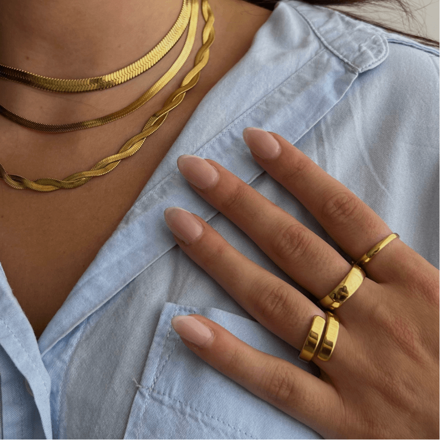 Person wearing multiple gold chain necklaces and gold rings with natural almond-shaped nails on light blue fabric