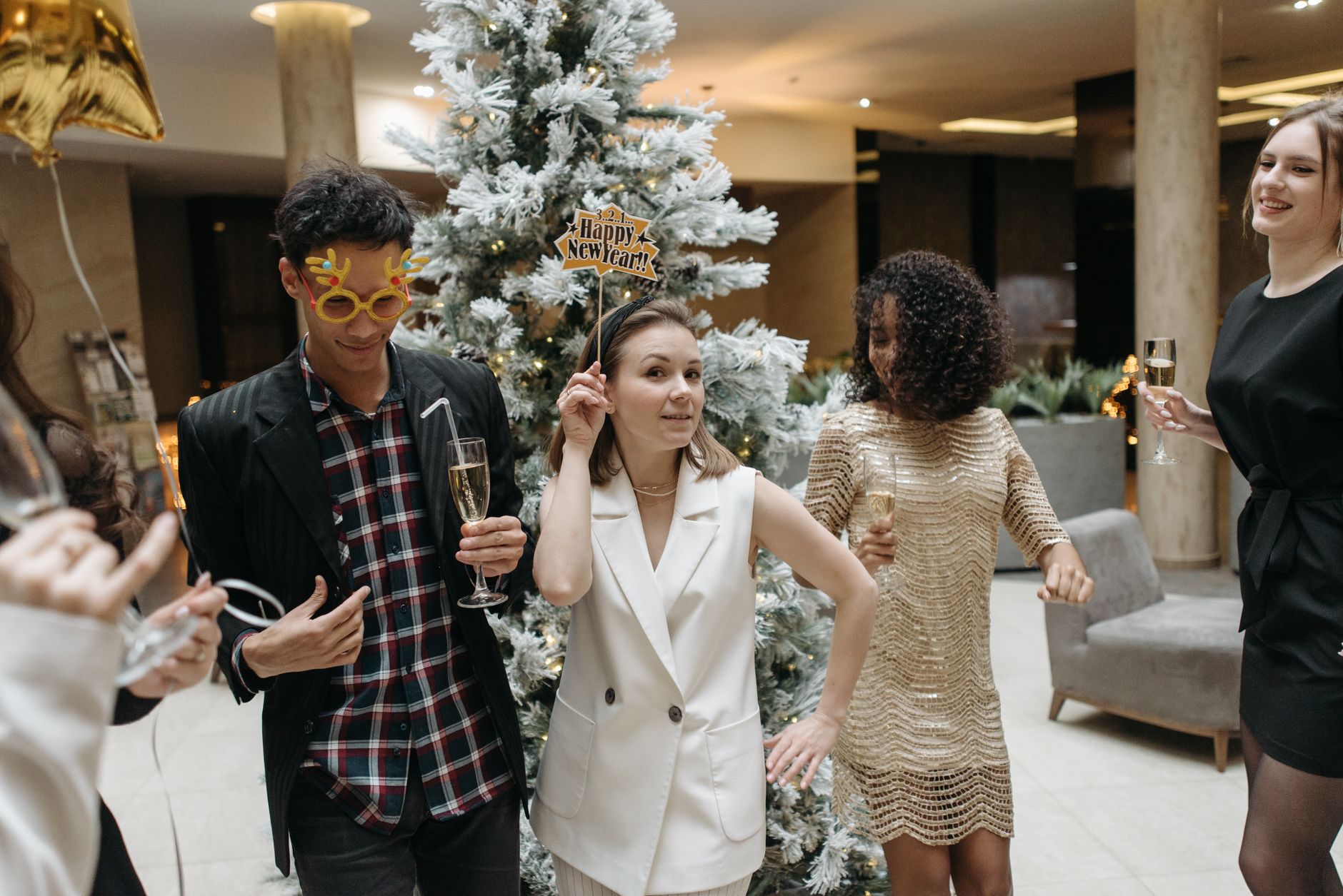 Group of people celebrating New Year's Eve indoors with a frosted Christmas tree, wearing party glasses and holding champagne glasses