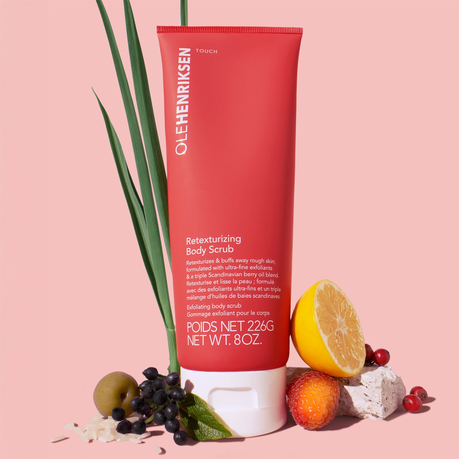 Red body scrub tube displayed on pink background surrounded by citrus, berries, and green leaves