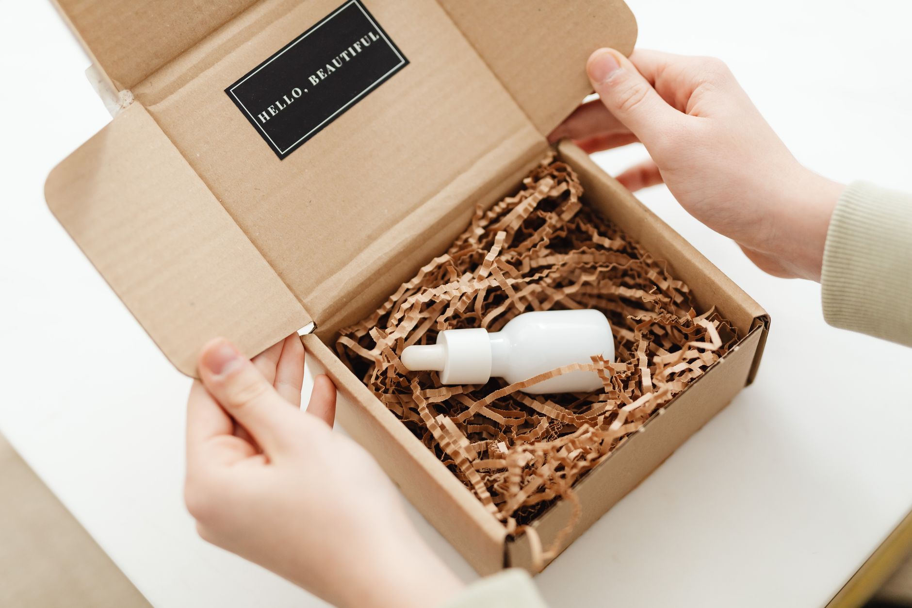 Hands opening cardboard box with brown crinkle fill and white beauty product bottles inside