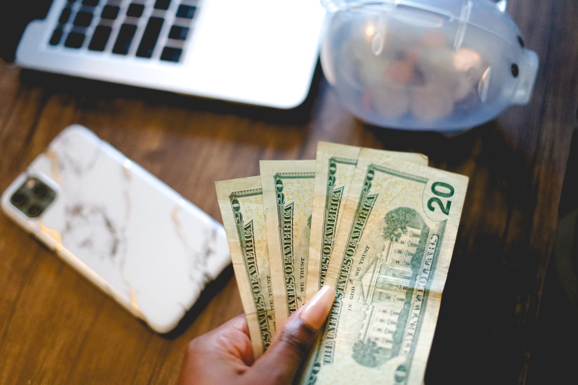 Hand holding five US twenty dollar bills fanned out on a wooden desk with a smartphone and water pitcher visible in the background