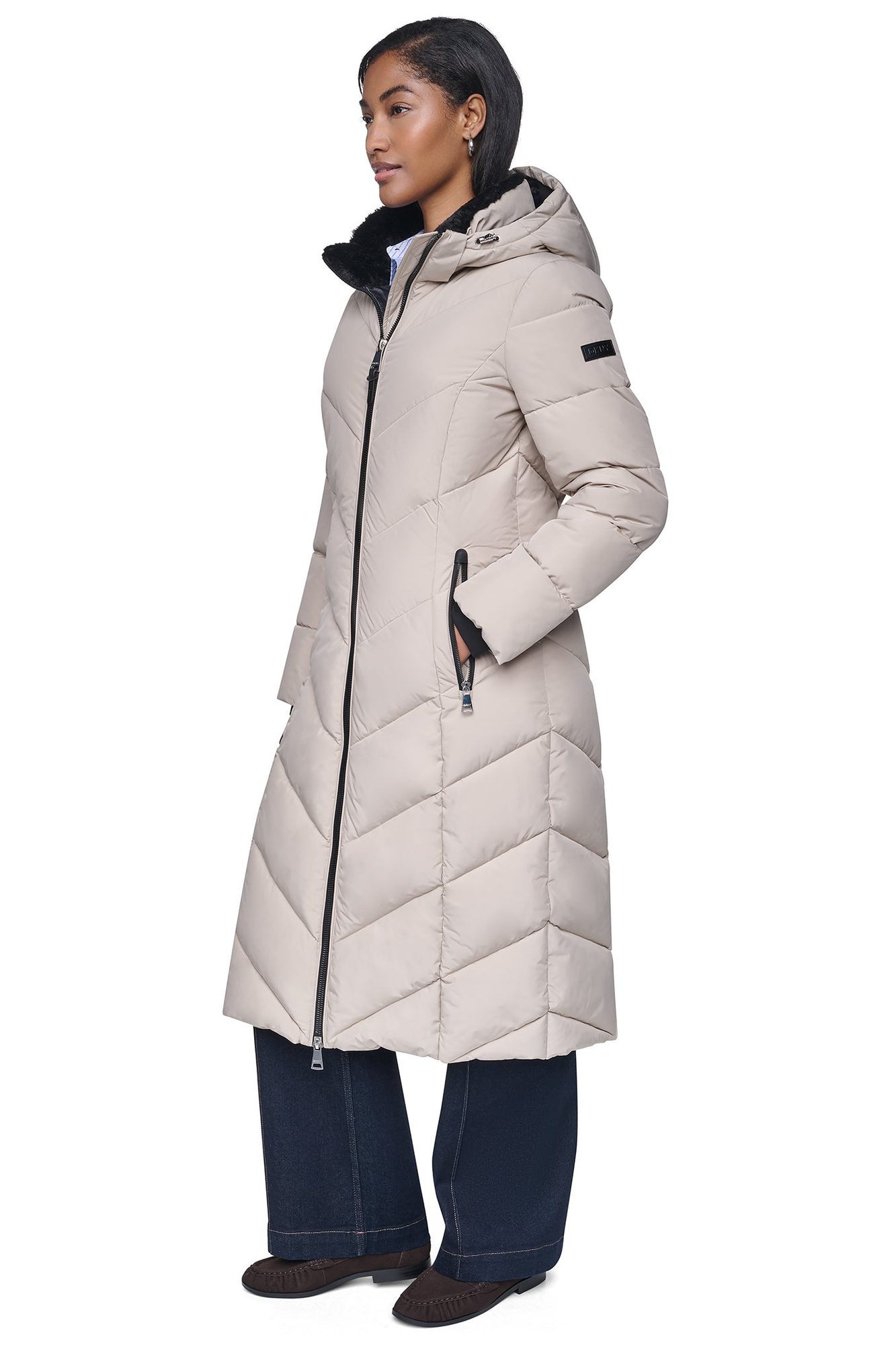 Woman wearing beige long puffer coat with black fur collar and hood over blue shirt and dark jeans