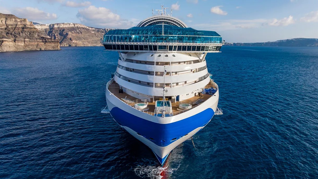 Modern cruise ship with blue and white hull on blue ocean near rocky coastline