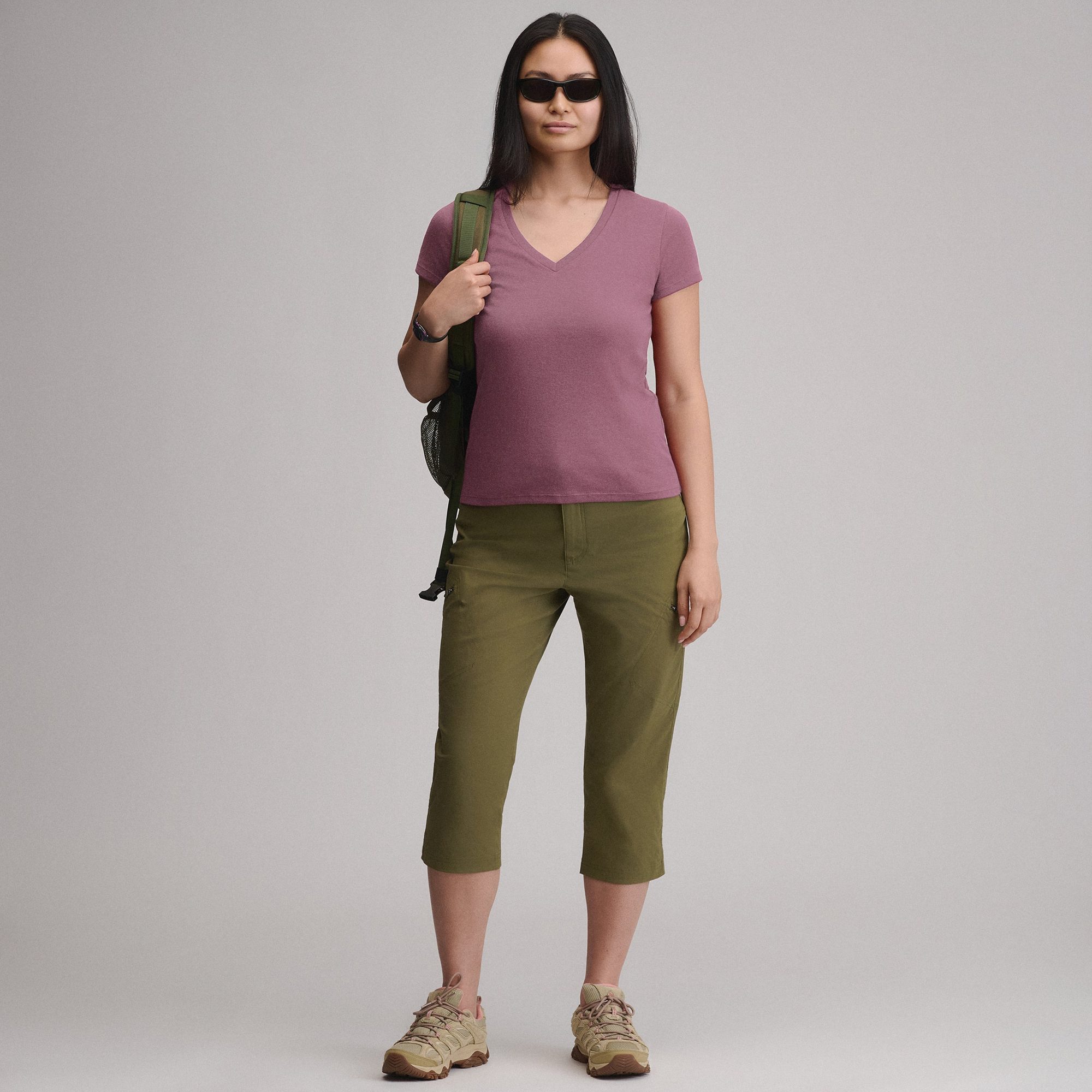 Woman wearing mauve v-neck t-shirt and olive green capris with backpack and hiking boots, full body view