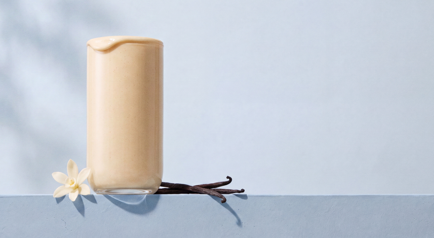 Glass of vanilla shake with vanilla bean pods and white flower on blue surface