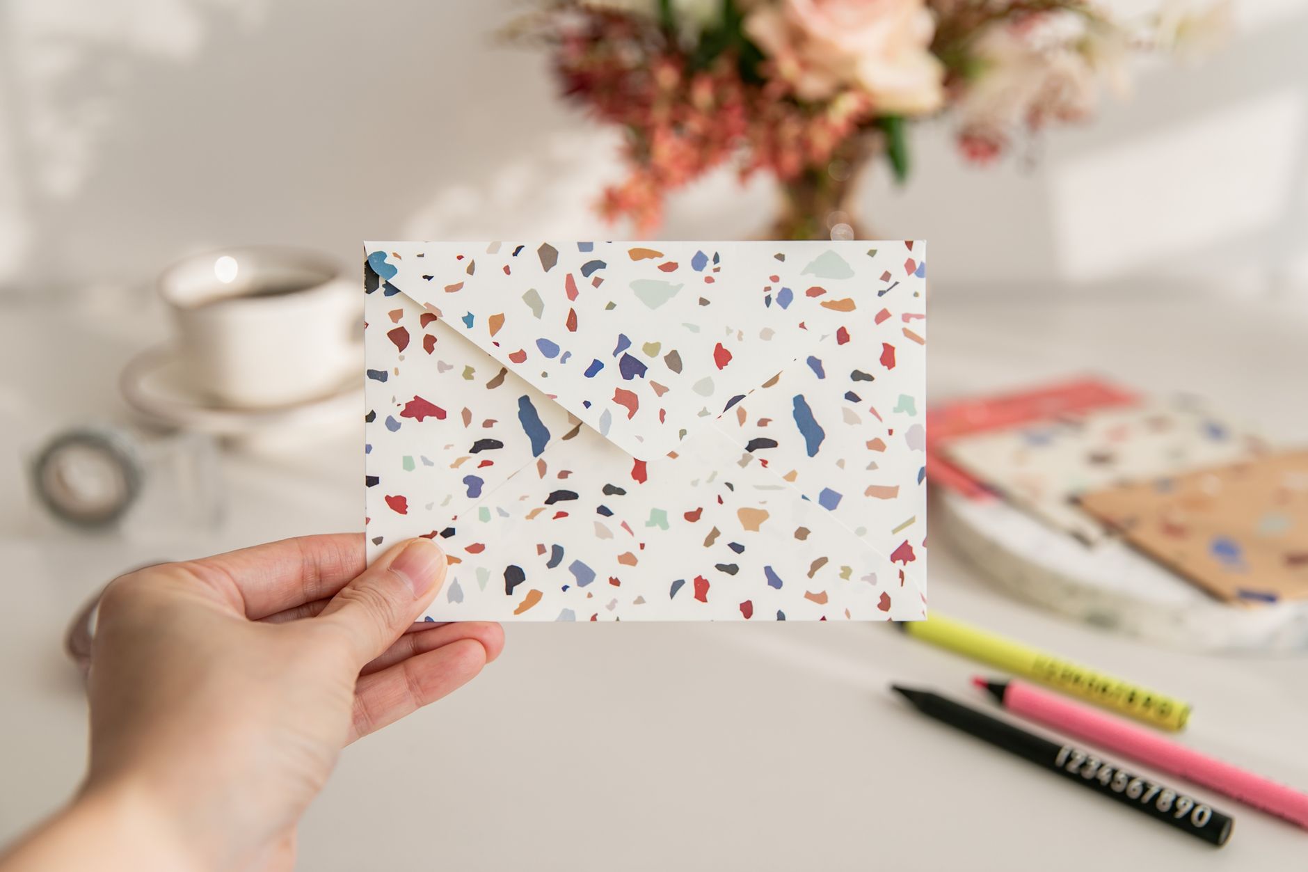 Hand holding colorful terrazzo-patterned envelope at desk with flowers, coffee cup, and colored pencils