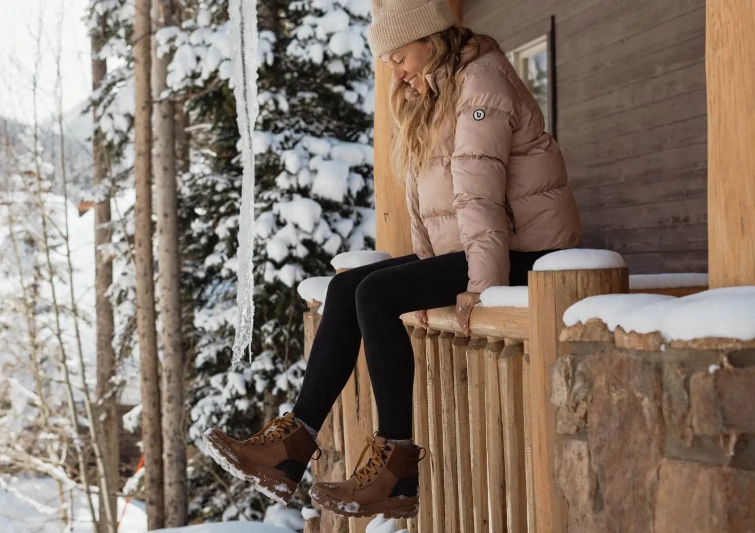 Woman in beige puffer jacket and black leggings sitting on a wooden railing in a snowy forest landscape