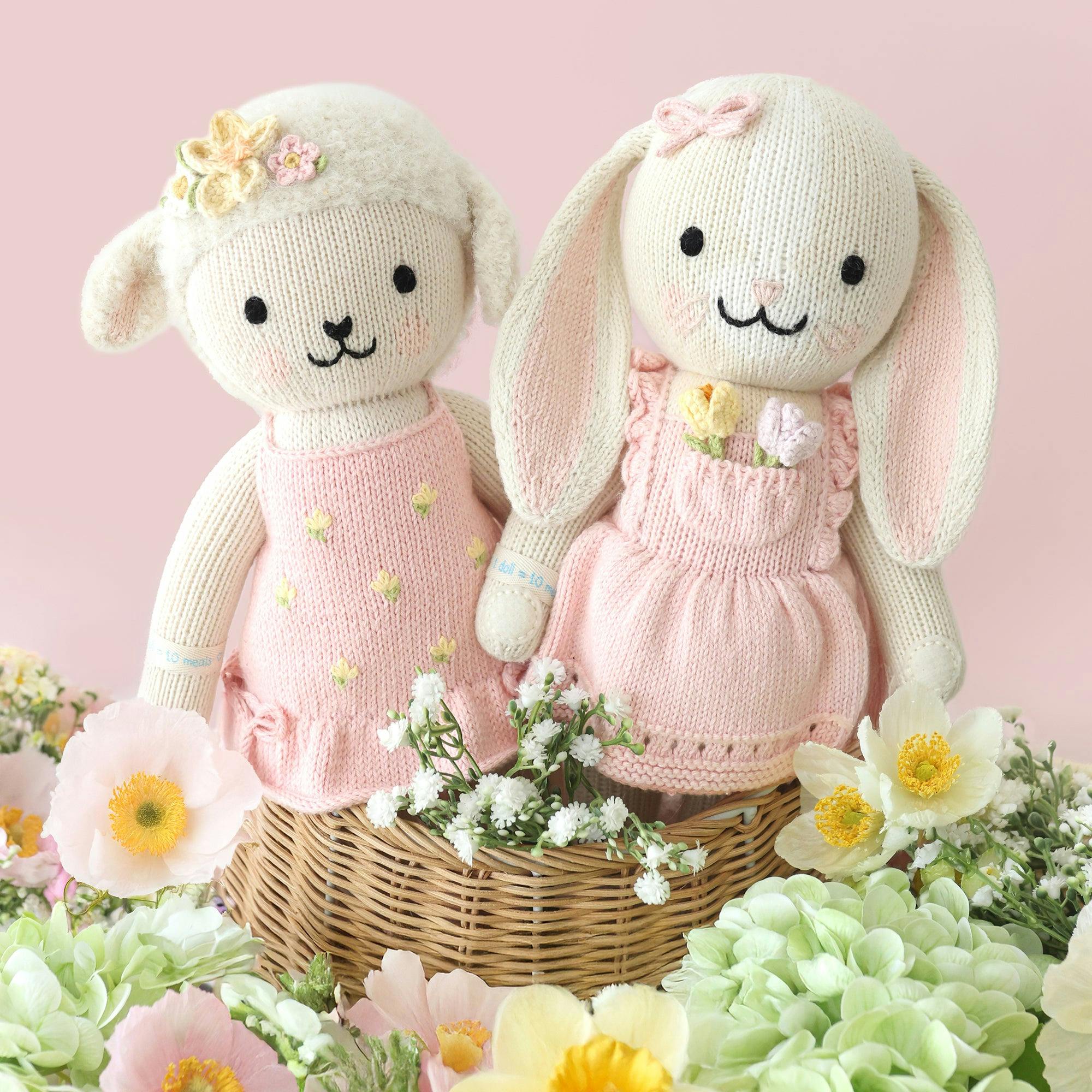 Two cream knit rabbit dolls in a wicker basket wearing pink dresses with embroidered flowers, surrounded by fresh daisies and greenery