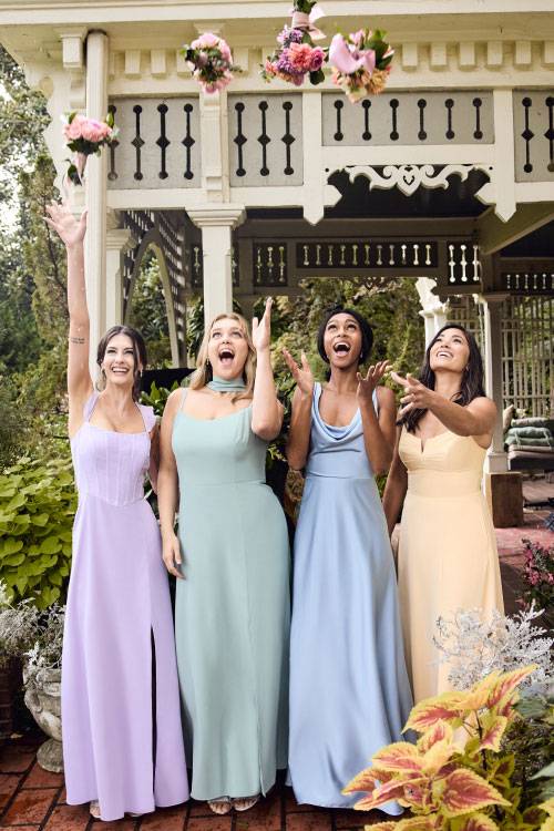Four women in pastel-colored bridesmaids dresses standing together under a white gazebo with hanging floral arrangements.