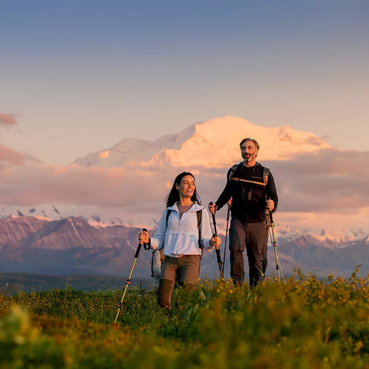 Couple hiking with trekking poles in alpine meadow with snow-capped mountain in background at sunset