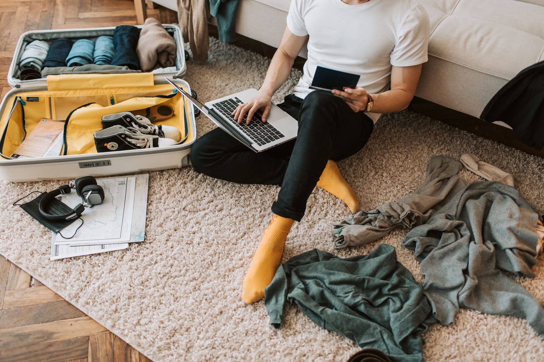 Woman sitting on carpet holding tablet and laptop, surrounded by open suitcase with packed clothes, headphones, and travel documents