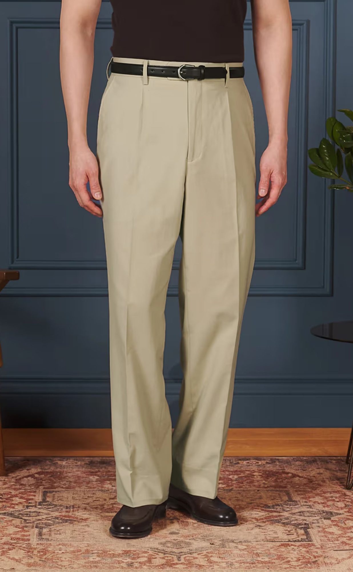 Man wearing cream pleated trousers with black belt and dark shoes, standing against blue paneled wall