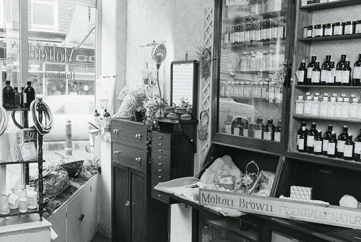 Black and white photograph of a vintage retail storefront interior with shelves of bottled products and branded signage