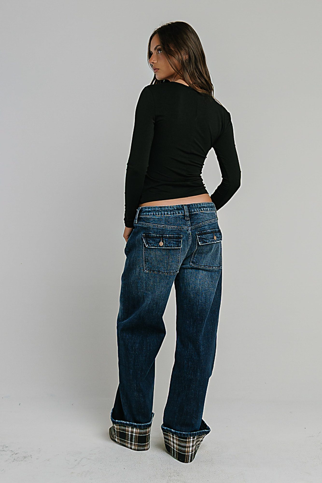 Woman in black long-sleeve crop top and dark blue jeans with plaid cuffs, viewed from behind against white background