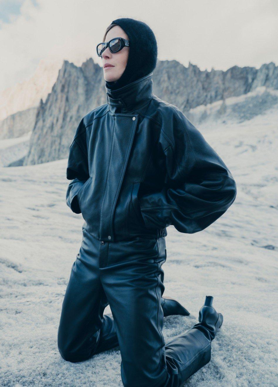 Person wearing black knit balaclava and blue winter jacket kneeling in snowy mountain landscape