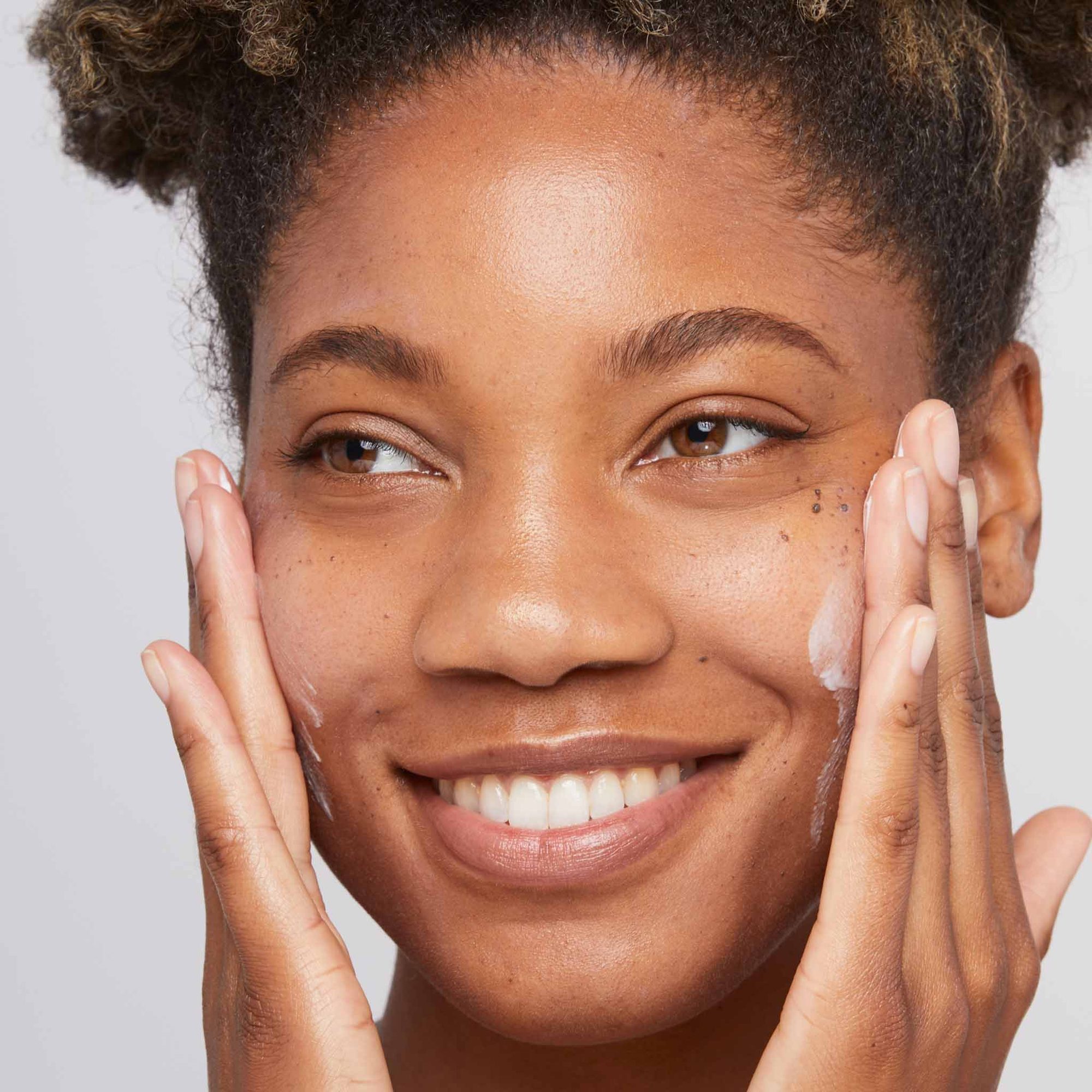 Woman with medium skin tone smiling while applying white cream to her cheeks with both hands