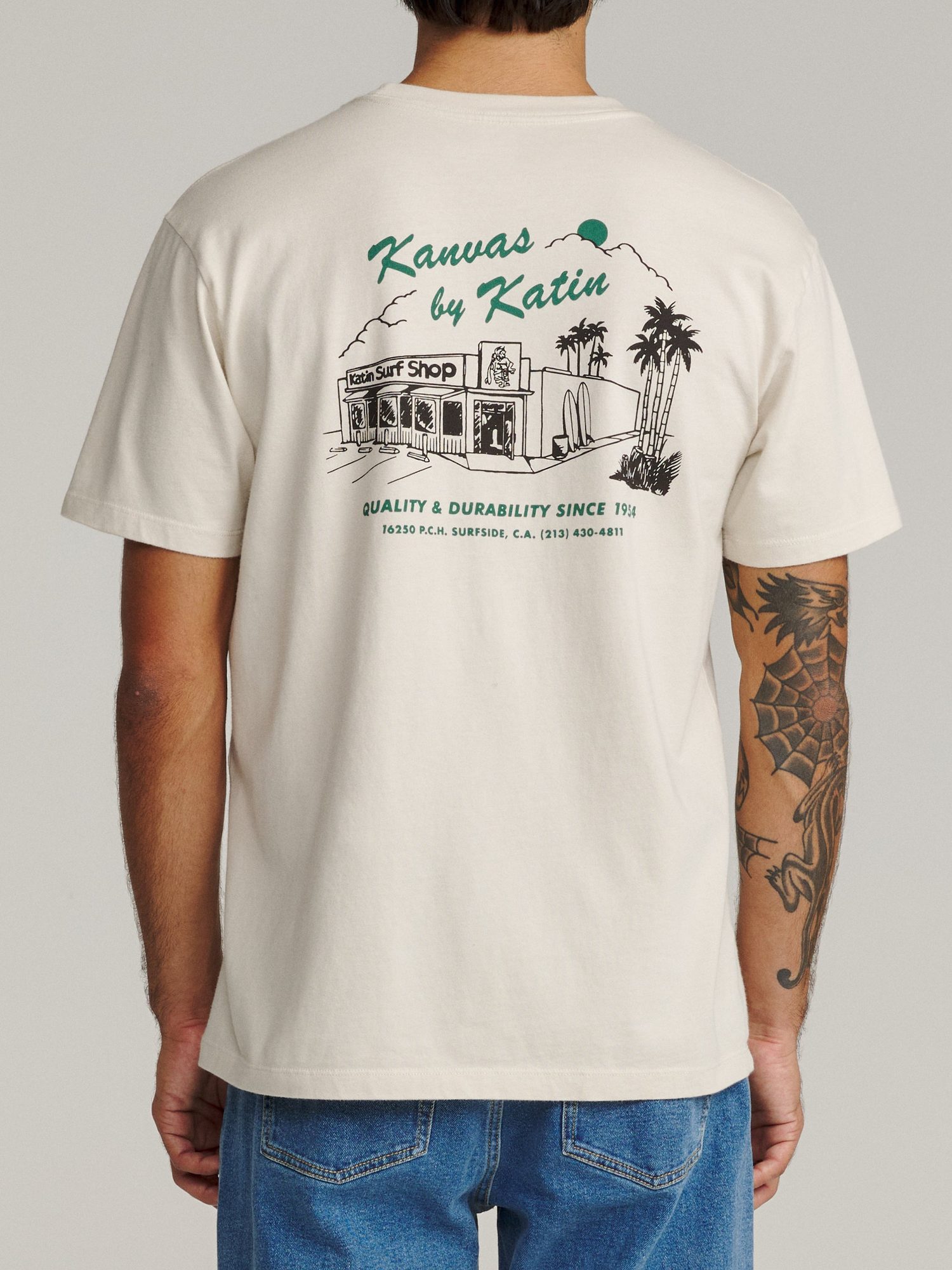 Back view of cream t-shirt with green 'Kanvas by Katin' text and surf shop illustration with palm trees