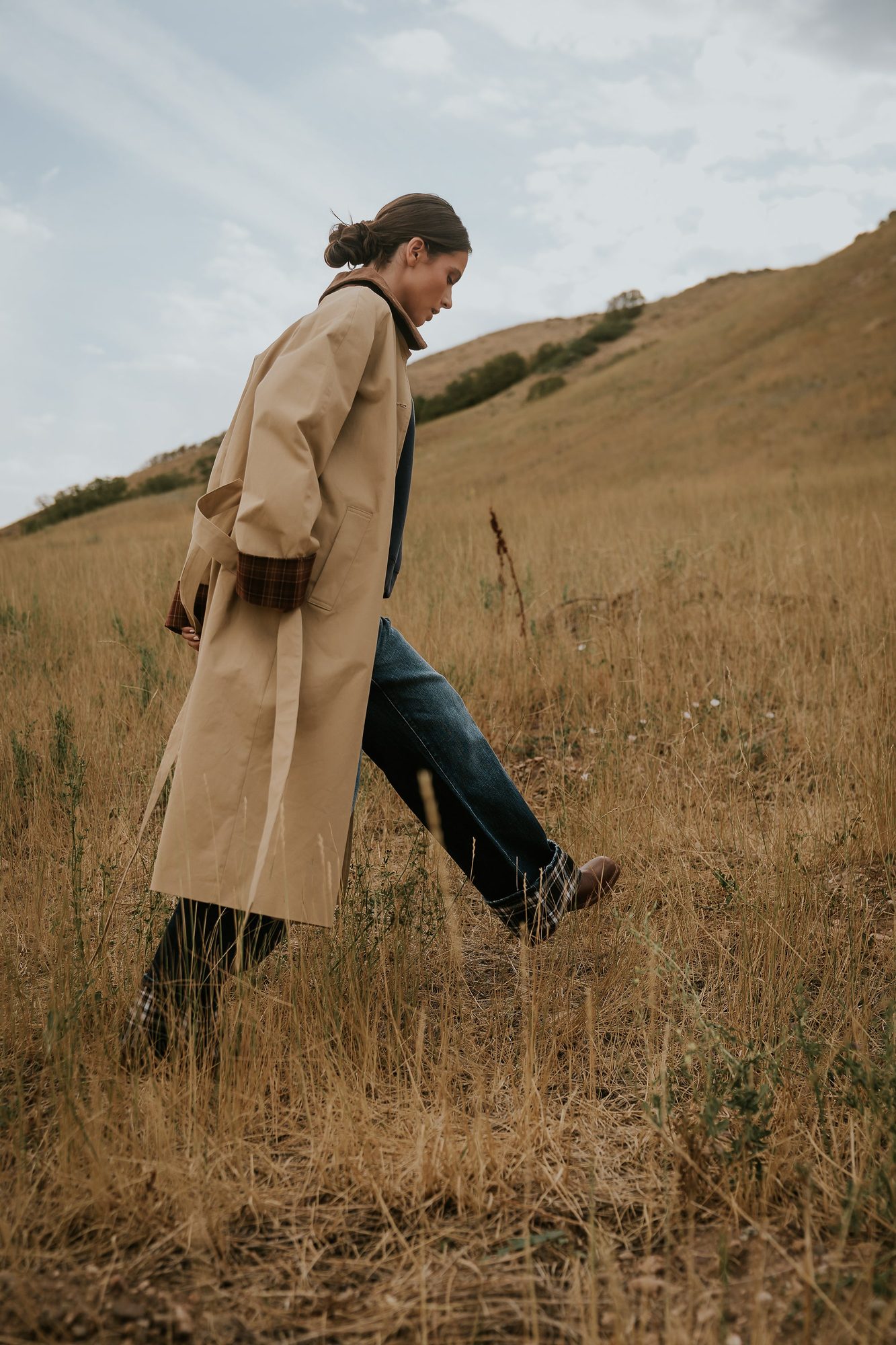 Woman in beige trench coat and dark jeans with plaid cuffs walking through dry field on hillside