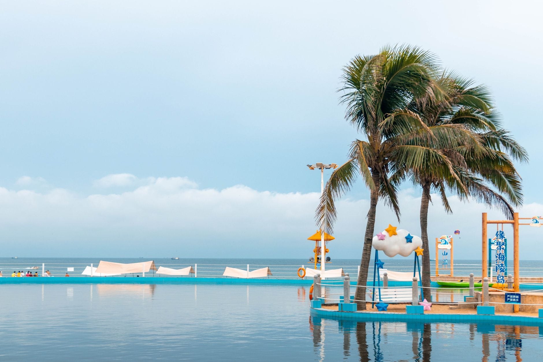 Beachfront pool with palm trees, blue water, and colorful play equipment overlooking calm ocean
