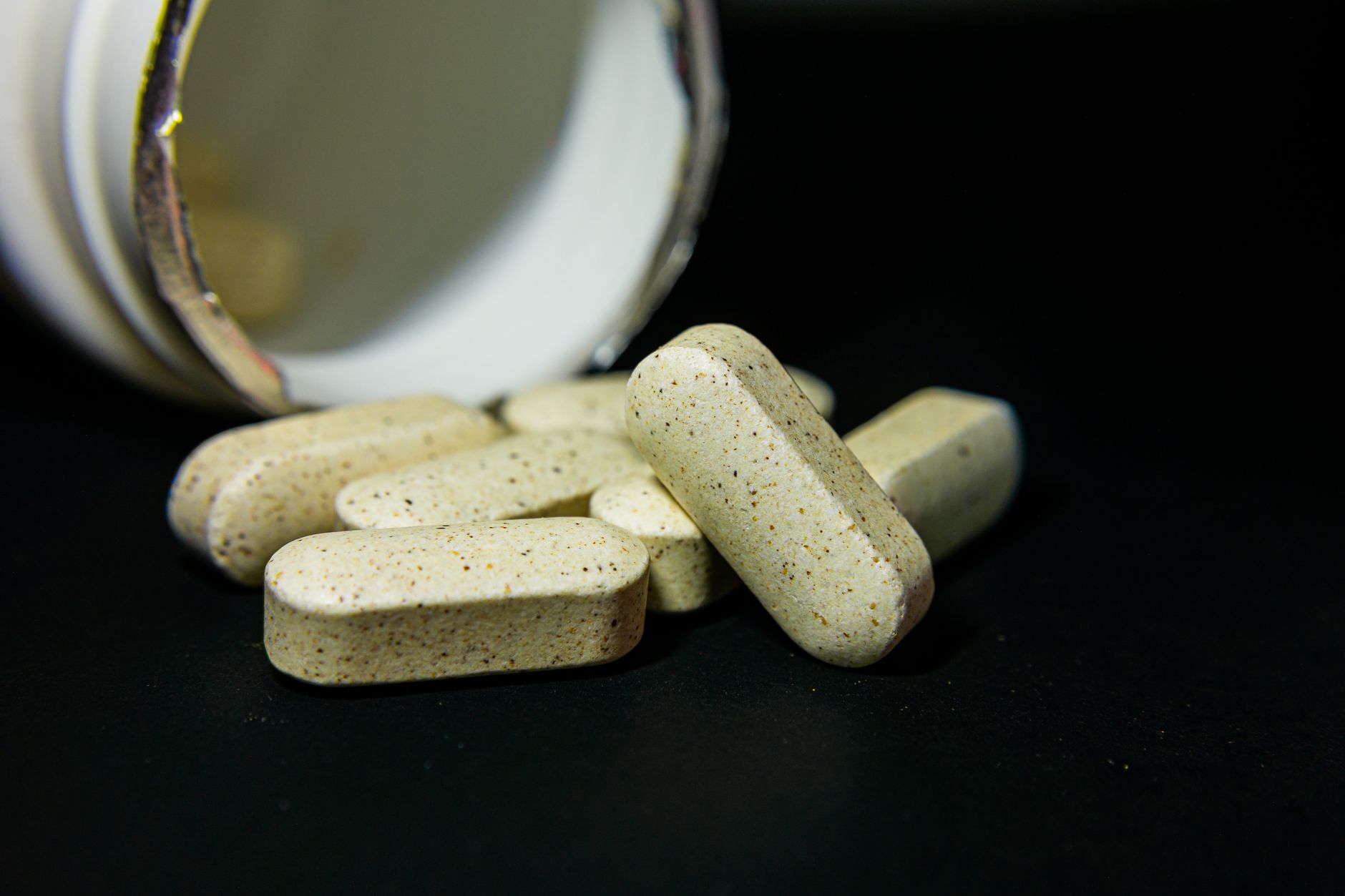 Tan and brown speckled capsule supplements spilling out of a white bottle on dark background