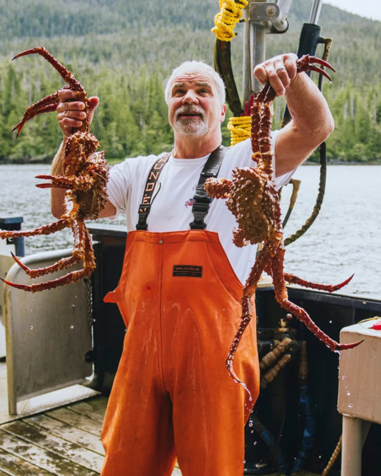 Fisherman holding two large red king crabs on boat with forest and lake behind him