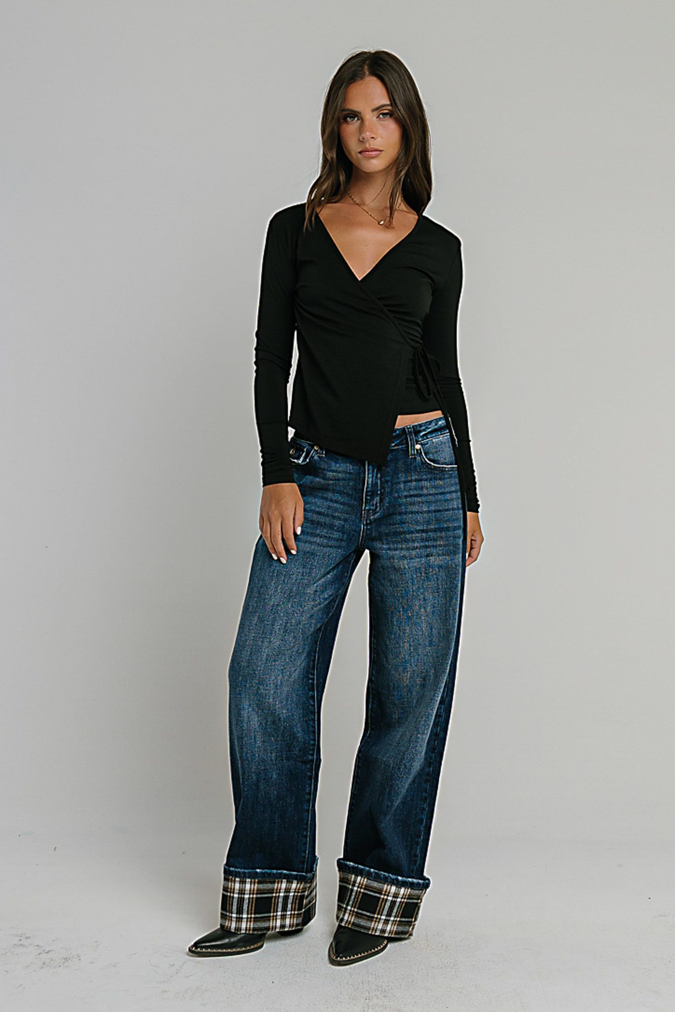 Woman wearing black wrap-style long-sleeve crop top and dark blue jeans with plaid cuffs, standing against white background