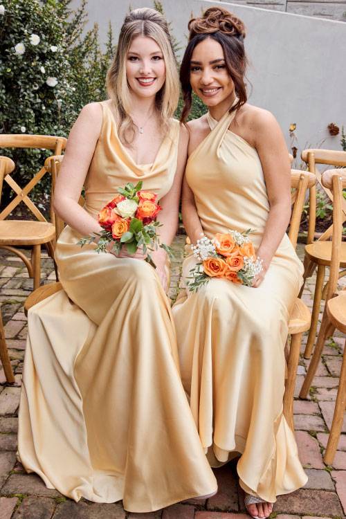 Two bridesmaids in champagne-colored satin dresses sitting outdoors holding orange and coral flower bouquets.
