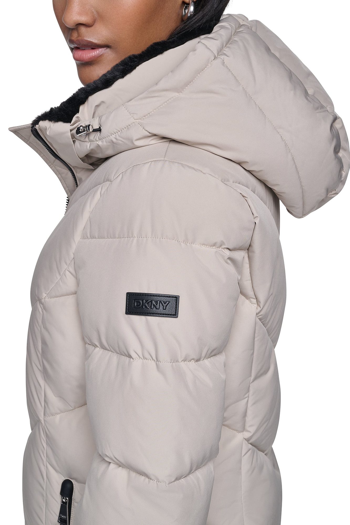 Close-up of beige puffer jacket showing quilted texture, black DKNY label patch, and black fur collar detail