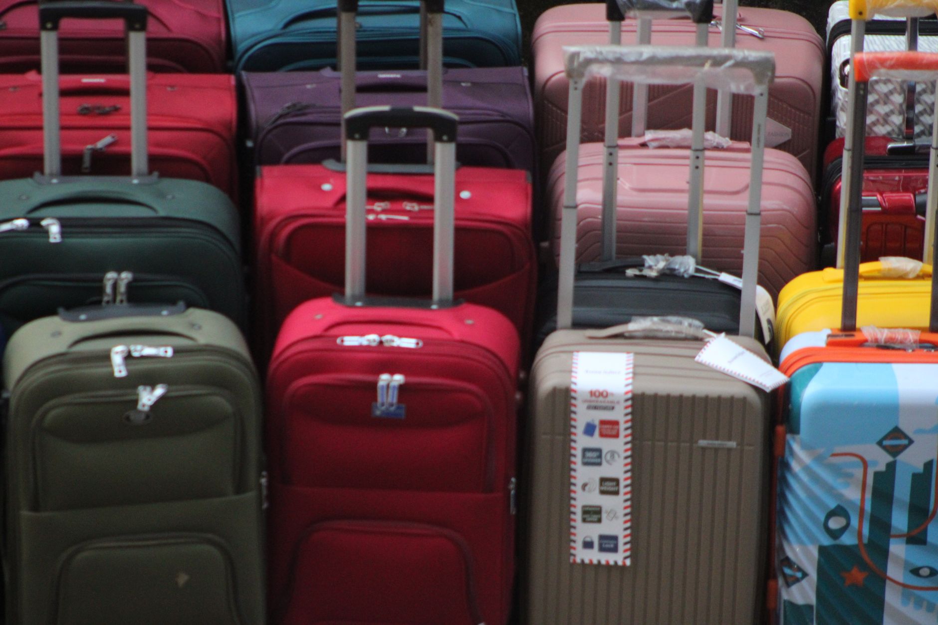Colorful hard-shell rolling suitcases stacked closely together in various colors including red, pink, blue, green, and yellow with retractable handles visible