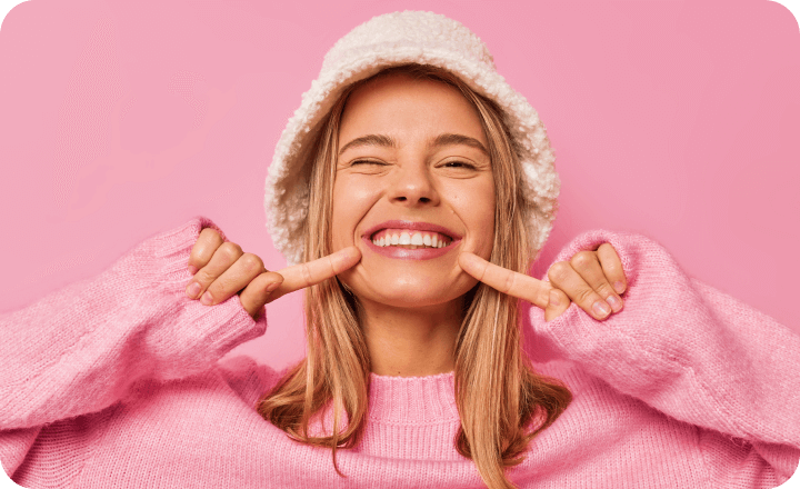 Young woman in pink sweater and white beanie pointing to her smiling teeth against pink background