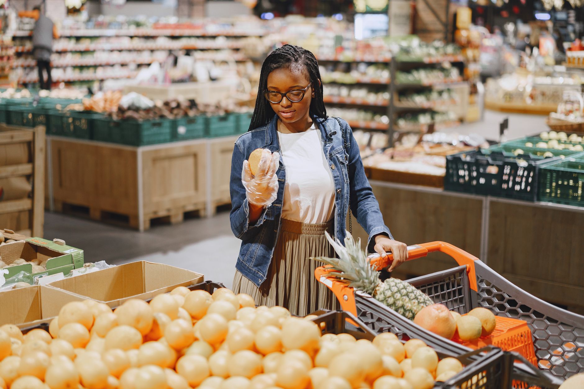 Woman in denim jacket holding grapefruit in produce warehouse with orange shopping basket full of groceries