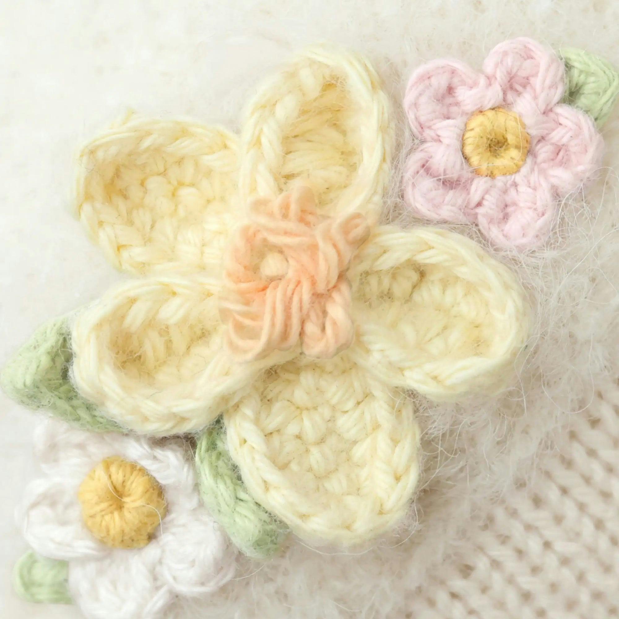 Embroidered knit flowers in cream, pink, and white with yellow centers on a textured background