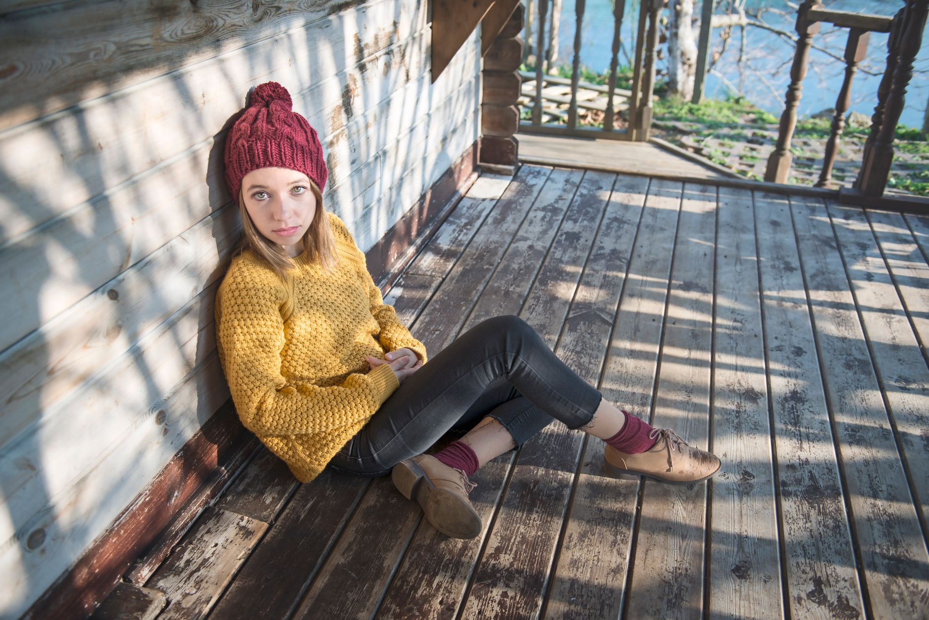 Girl sitting on wooden deck wearing yellow sweater, burgundy beanie, black leggings, and tan boots